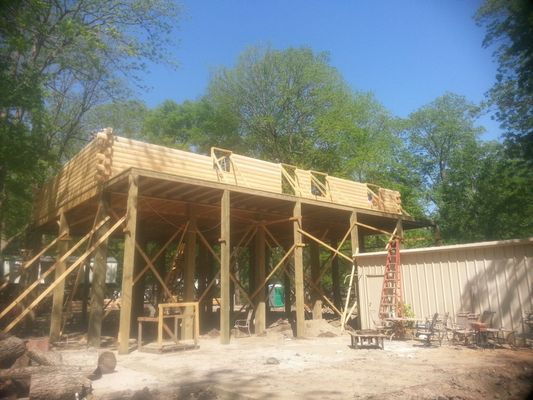 Wooden structure under construction, supported by posts, in a wooded outdoor setting.