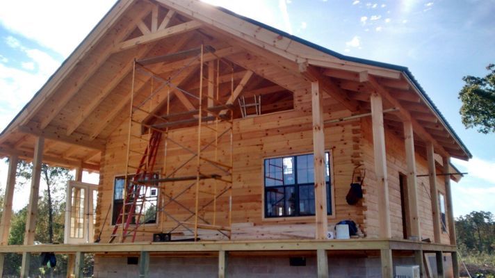 A wooden house under construction with exposed beams, a porch, windows, and scaffolding.