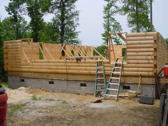 Log cabin under construction, logs stacked on a concrete foundation, with ladders and tools.