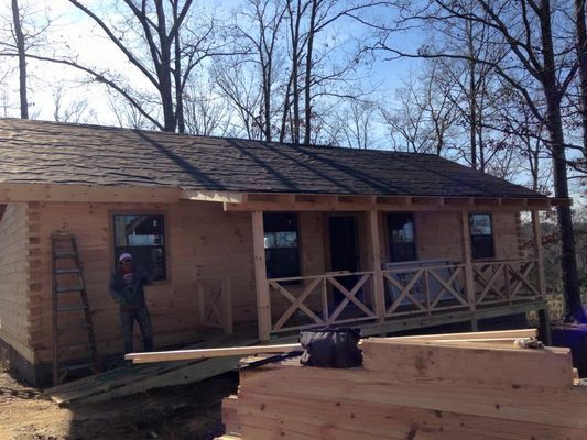 Wooden cabin under construction with a porch and a person standing outside.
