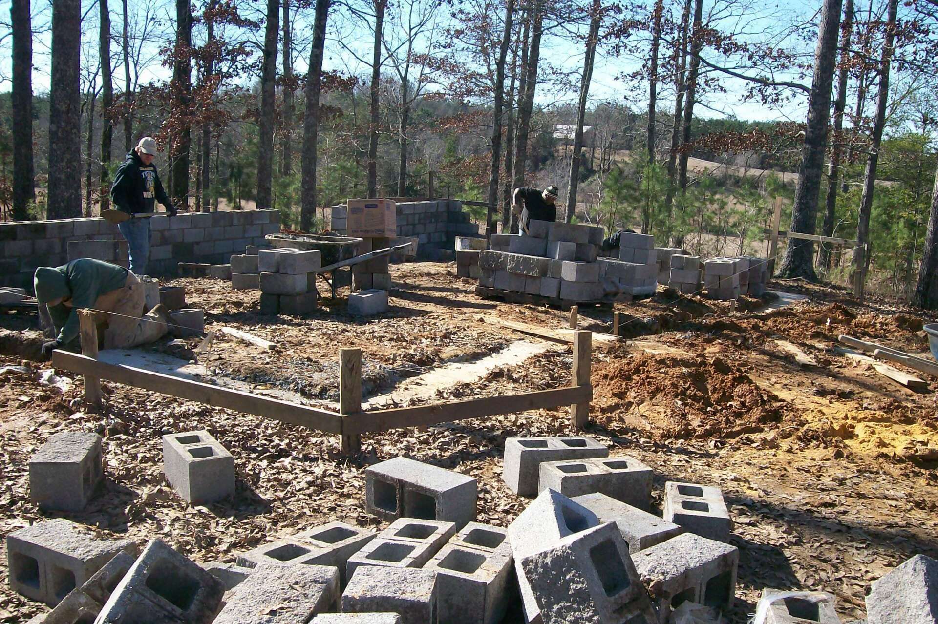 Construction site with cinder blocks, workers, and wooded background.