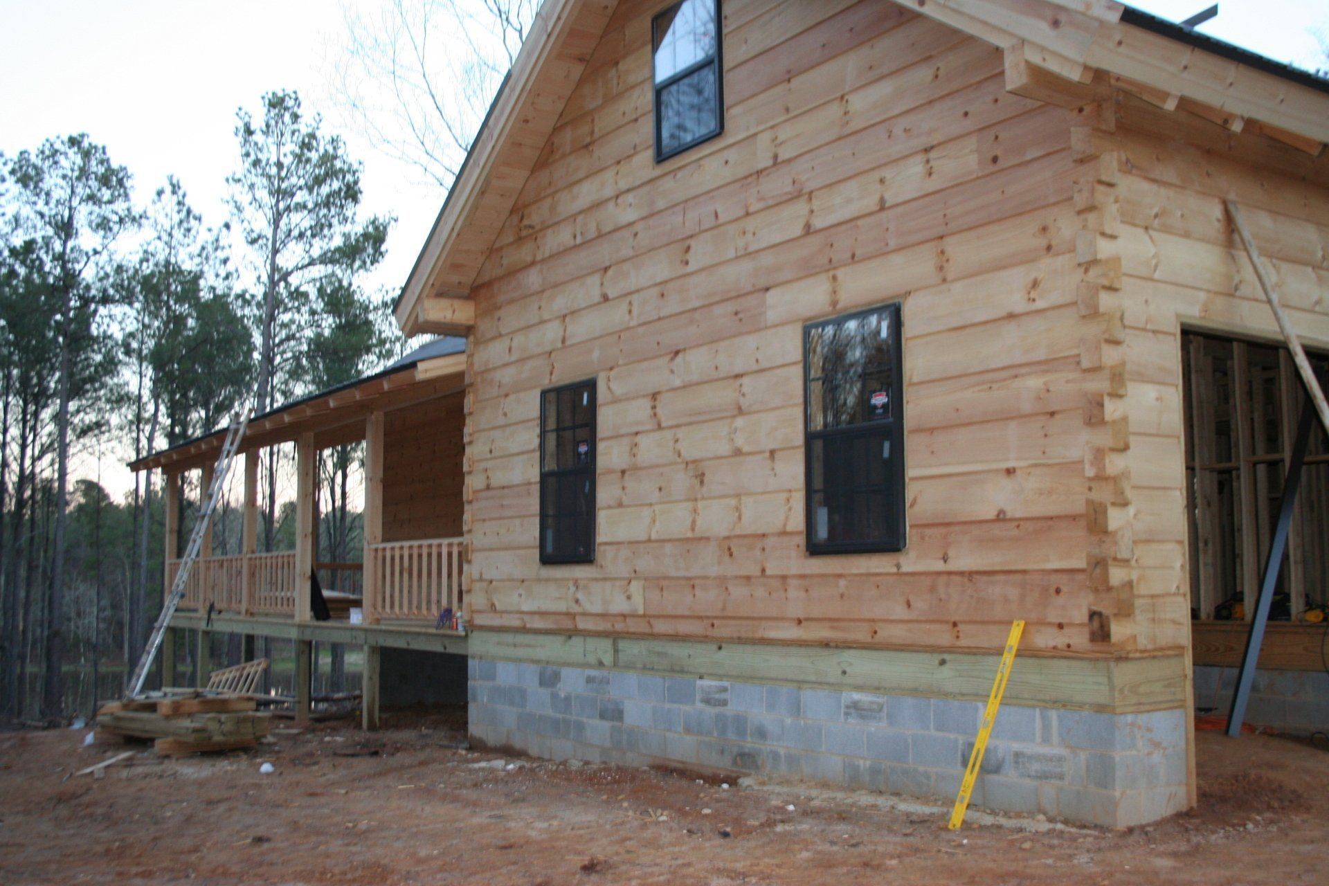 Log cabin under construction with wood siding, windows, and porch, on a concrete foundation.