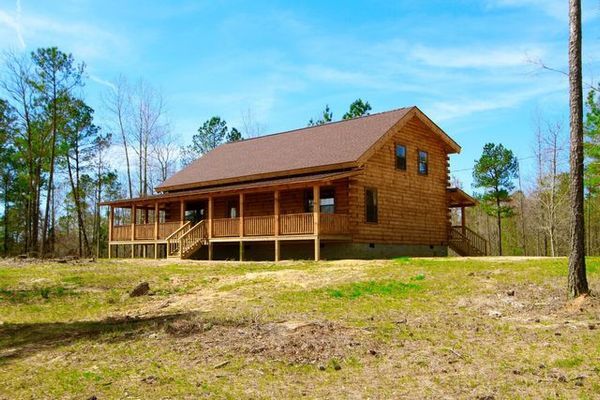 Wooden log cabin with a porch and brown roof, set in a grassy, wooded area under a blue sky.