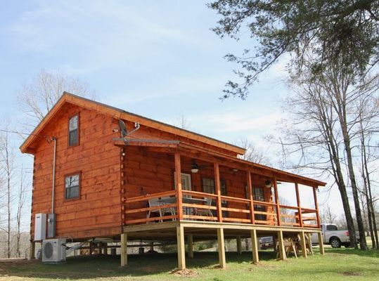 Wooden cabin with a covered porch on stilts, set in a grassy outdoor area.