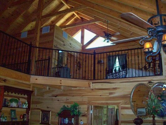 Interior of a log cabin with a loft and black railing; visible beams, ceiling fan, and decorative chandelier.