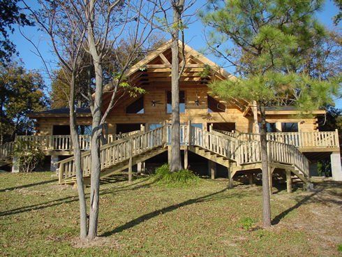 Wooden cabin with a wrap-around deck, accessed by wooden stairs, surrounded by trees on a grassy hill.