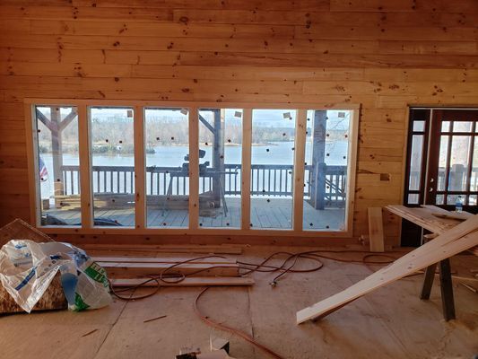Interior of a wooden cabin with large windows overlooking a lake and deck during construction.