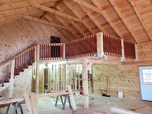 Interior view of wooden building under construction, showing a loft with stairs and railing, and exposed studs.