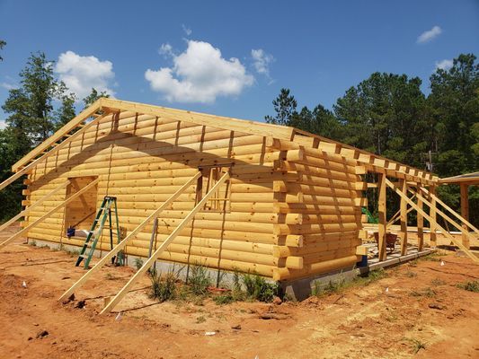 Log cabin under construction with wood frame roof and support beams, outdoors on a sunny day.
