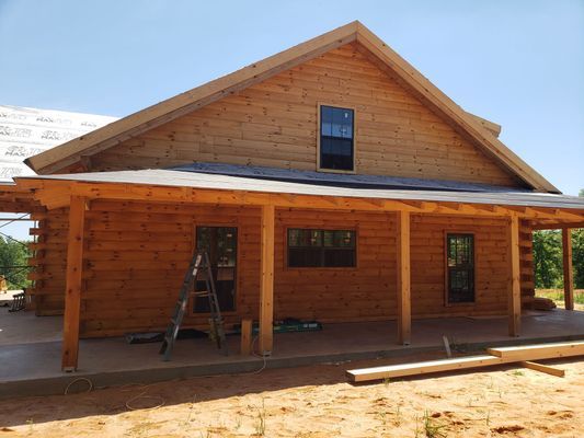 Log cabin under construction with a wraparound porch, blue sky.