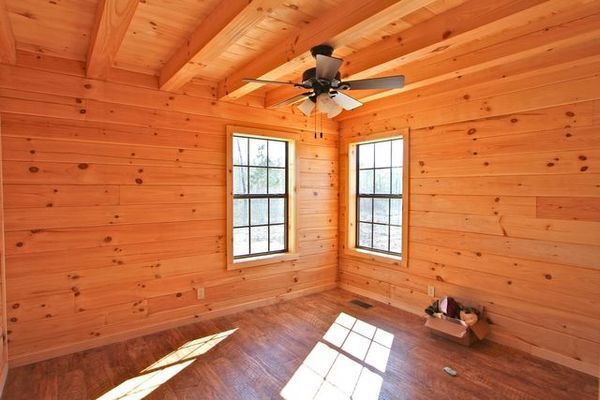 Wooden interior of a room with two windows, ceiling fan, and light shining on the wood floor.