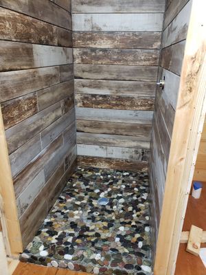 Shower stall with wood-look tile walls and pebble floor; unfinished wooden framing visible.