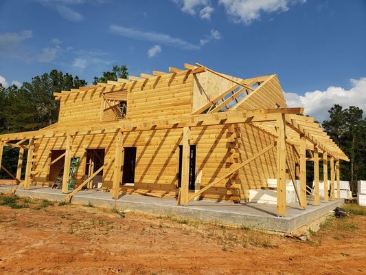 Wooden house under construction on a concrete foundation, surrounded by dirt and trees, sunny day.