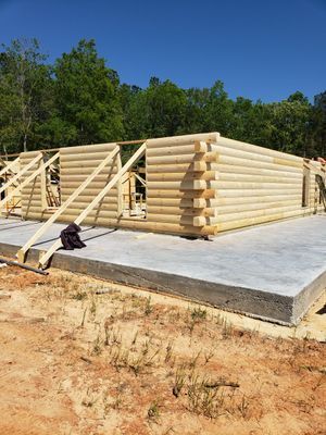 Log cabin under construction on a concrete foundation, with wooden logs stacked. Trees and blue sky in the background.