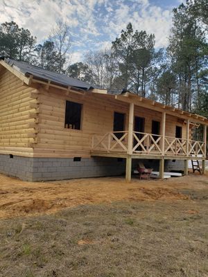 Log cabin under construction with porch and open windows, surrounded by trees and dirt.