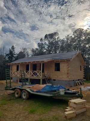 Log cabin under construction with porch, trailer, and lumber outside. 
