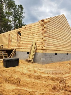 Log cabin under construction with stacked wooden logs on a cinder block foundation, set in a dirt lot.
