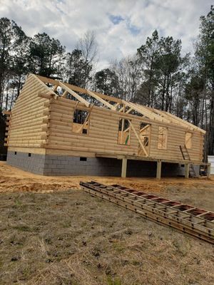 Log cabin under construction; wooden walls on concrete foundation, roof framing, ladder.
