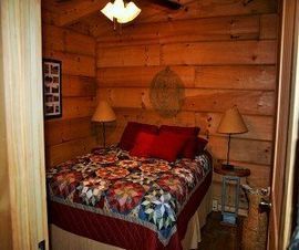 Cozy bedroom with log walls, floral quilt, bedside lamps, and a small woven wall hanging.