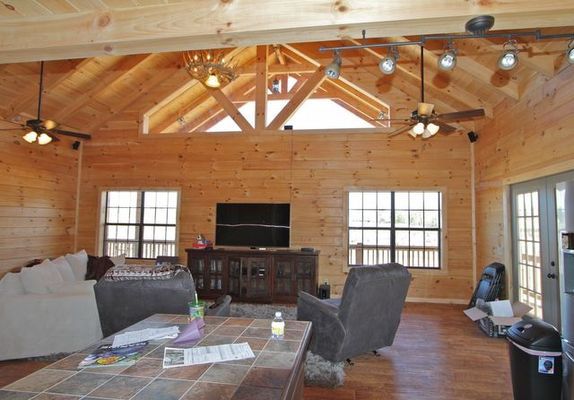 Interior of a log cabin living room with wooden walls, high ceilings, ceiling fans, windows, TV, and furniture.