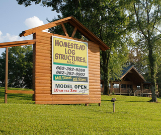 Sign for Homestead Log Structures, LLC, next to a log cabin model on a grassy lawn.