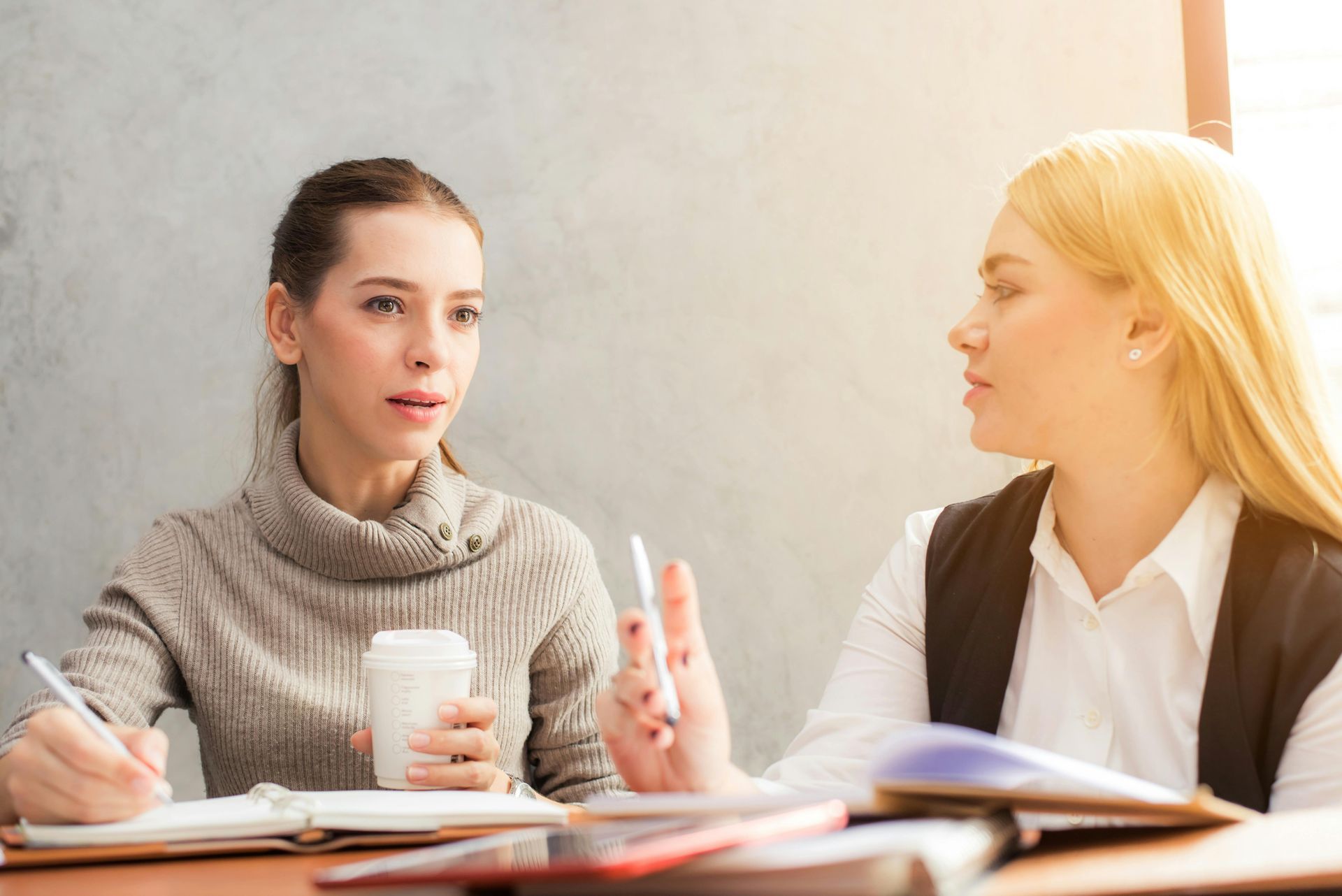 Two women in an office setting, one writing and holding coffee, the other gesturing with a pen, engaged in conversation.