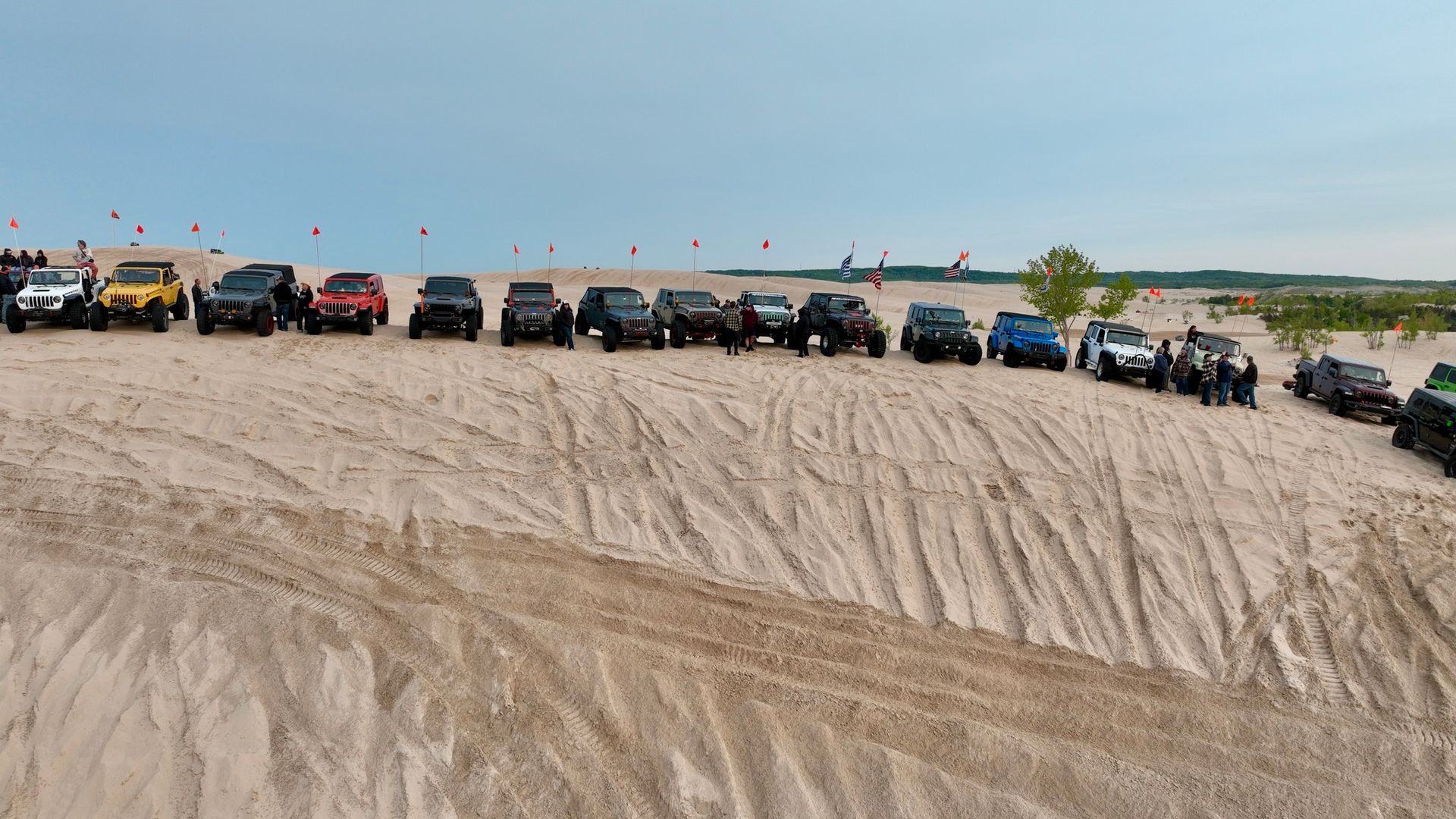 A line of Jeeps parked on a sandy dune, with people standing around; the sky is blue.