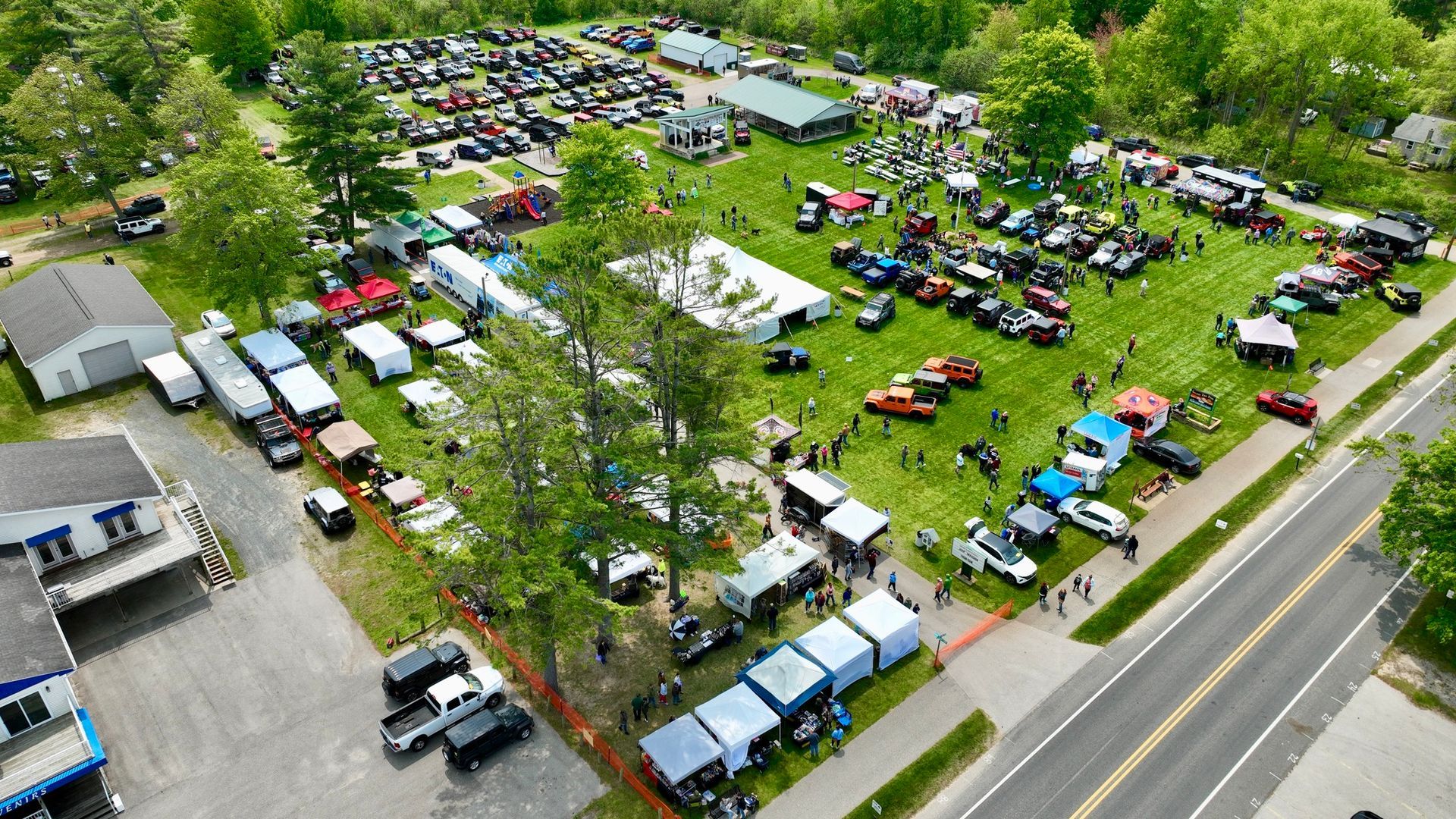 Aerial view of a car show with numerous vehicles parked on grass, vendors, and spectators.