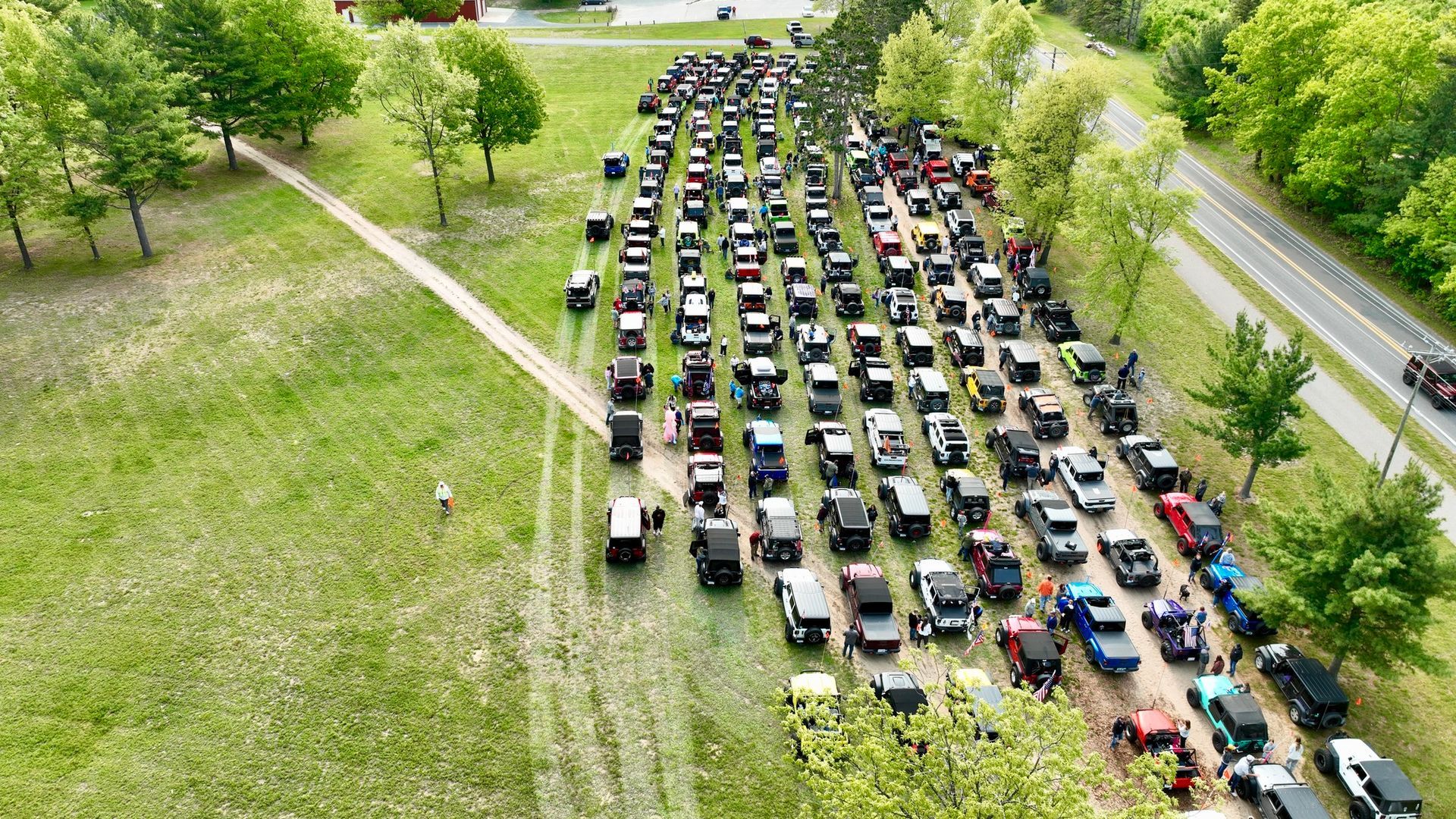 An aerial view of a large gathering of Jeeps parked in a field next to a road, some people are present.
