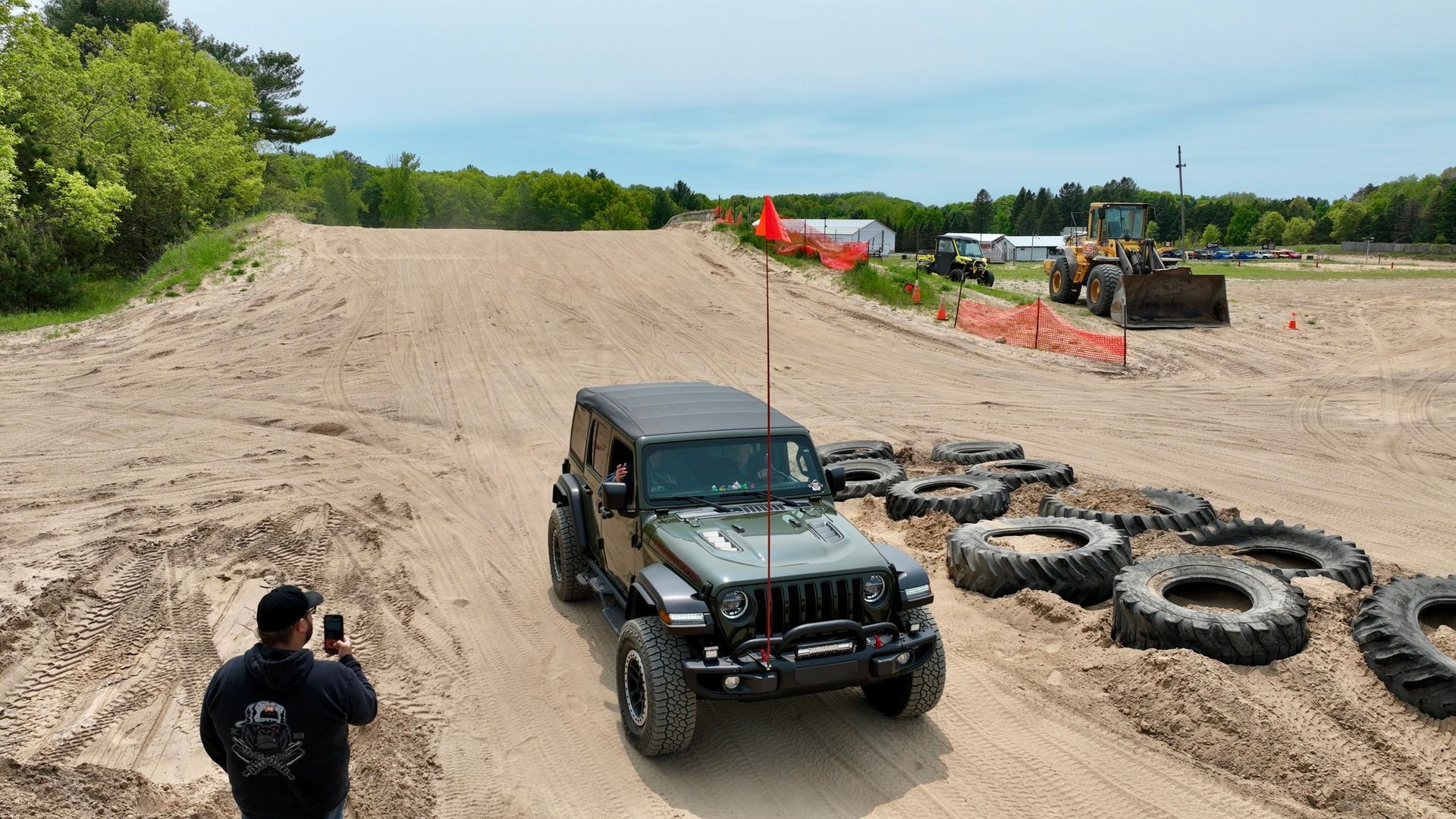 Olive green Jeep climbs a sandy hill, person taking photo. Tires and construction equipment visible.