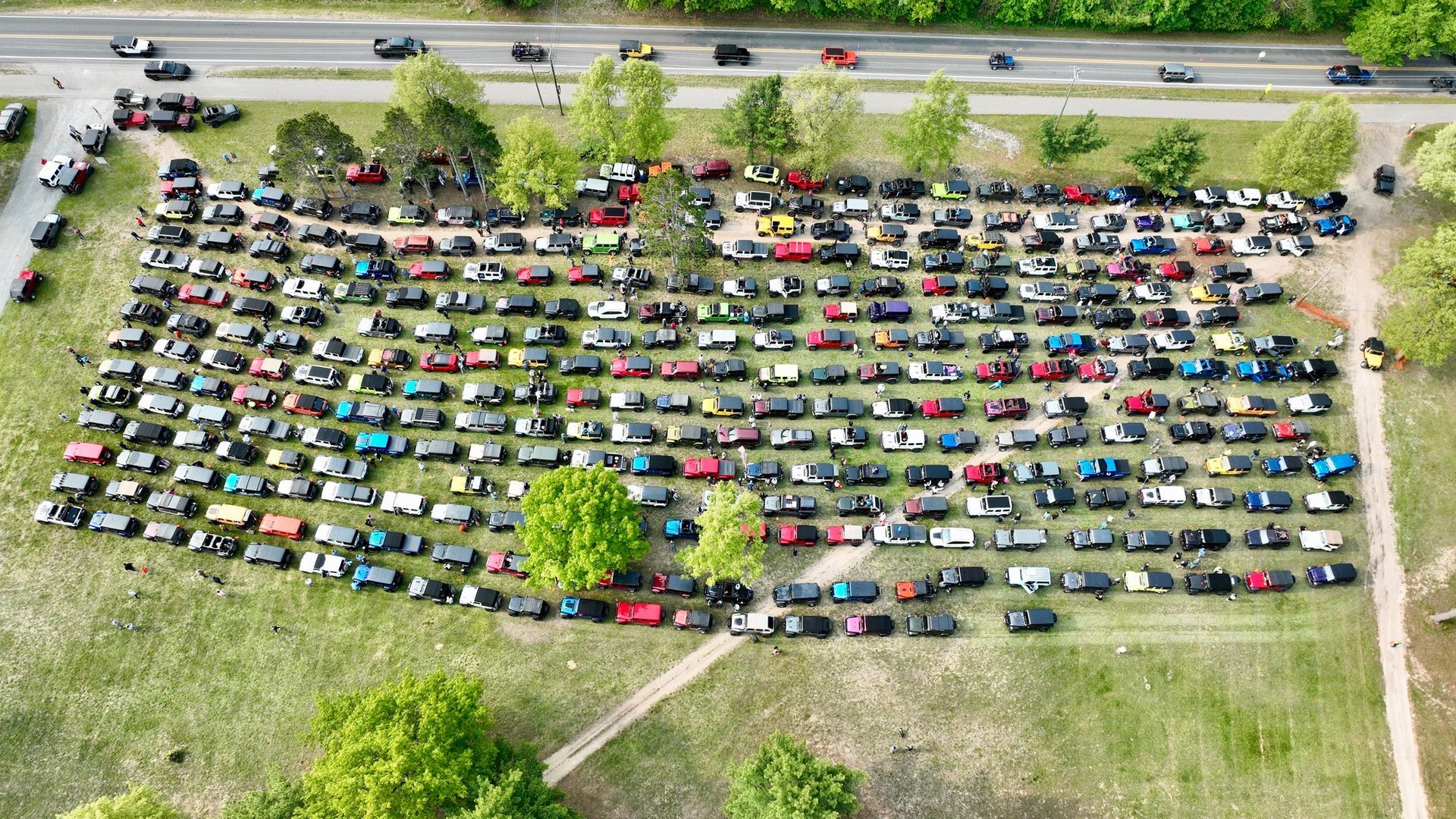Overhead view of a large gathering of cars in a grassy field next to a road, mostly dark colors.