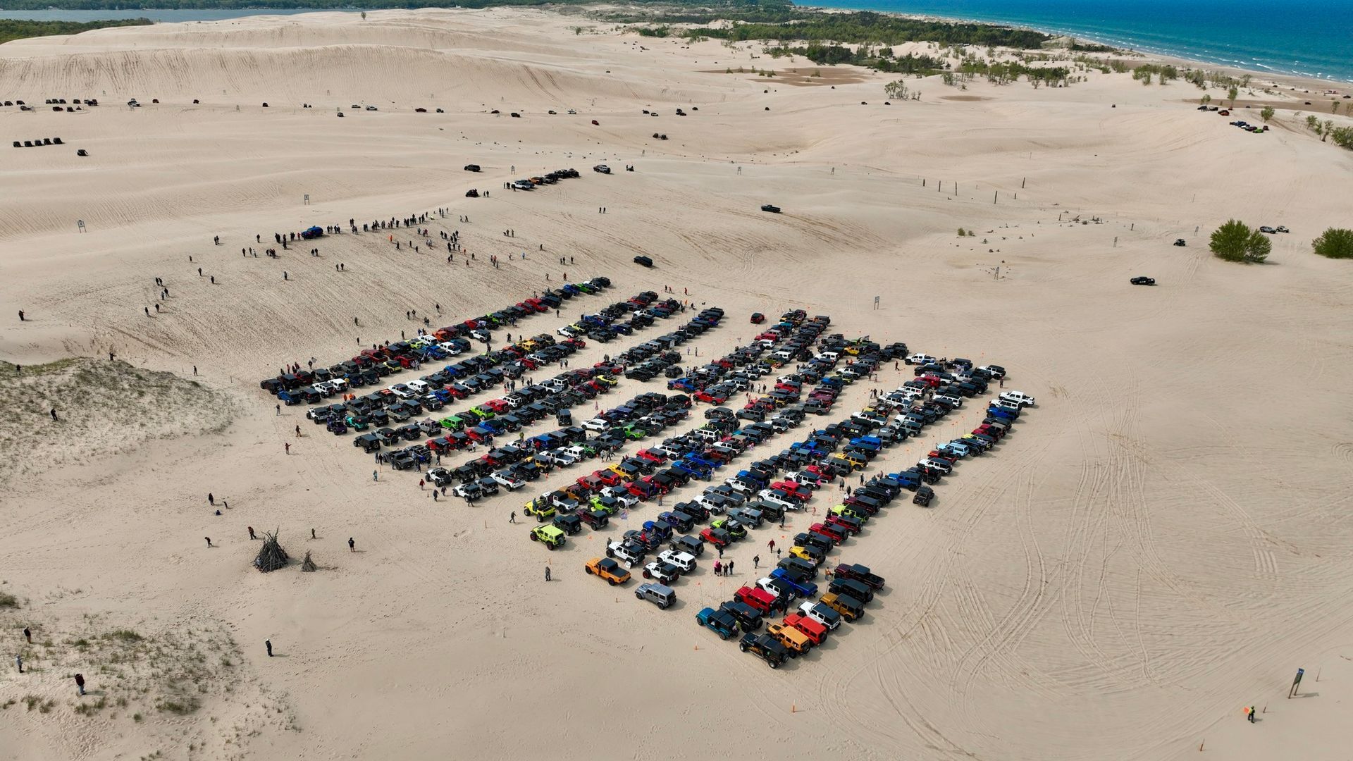 Cars parked in neat rows on a sandy dune. People stand nearby, and trees and water are in the background.