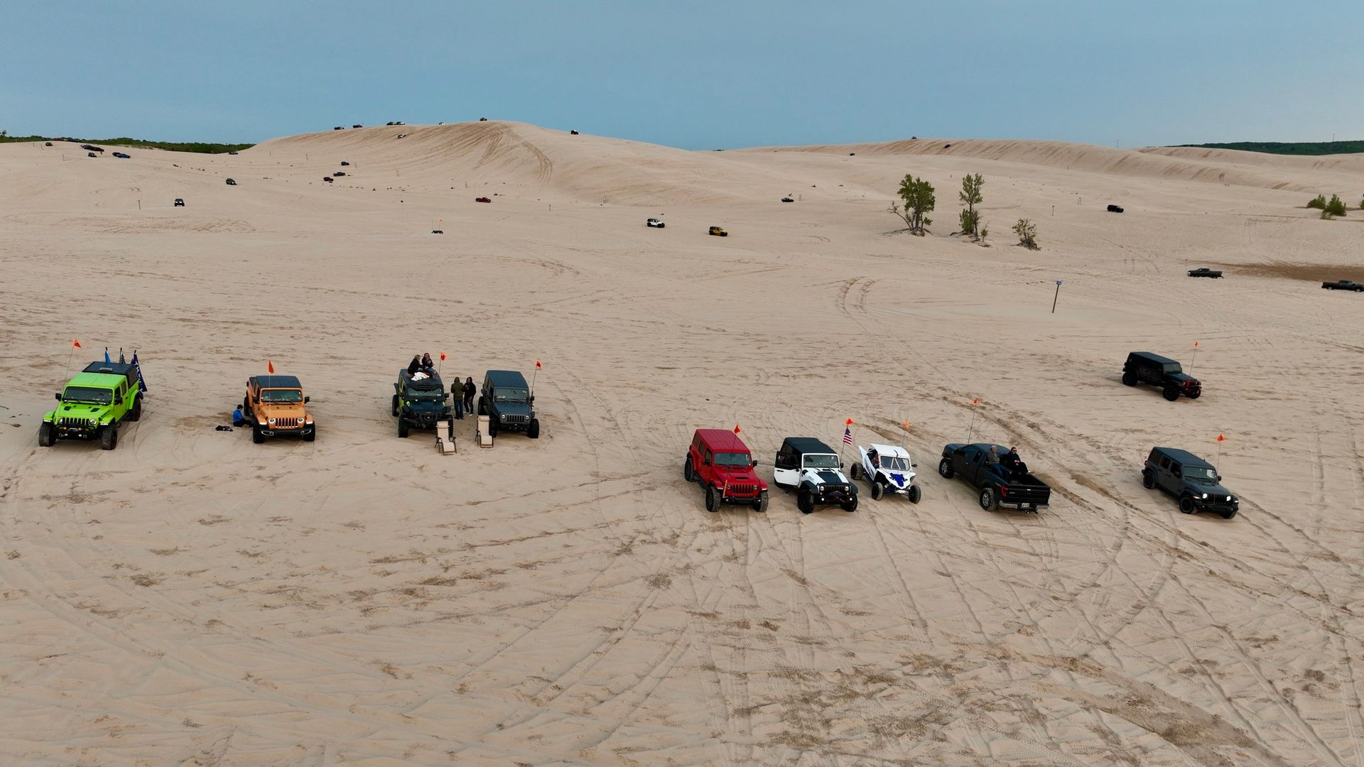 Off-road vehicles parked on a sandy dune. Several people stand near the vehicles. Overcast sky.