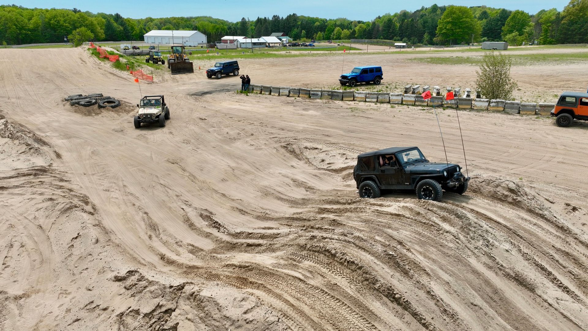 Jeeps driving on a sandy off-road course with spectators and buildings in the background.