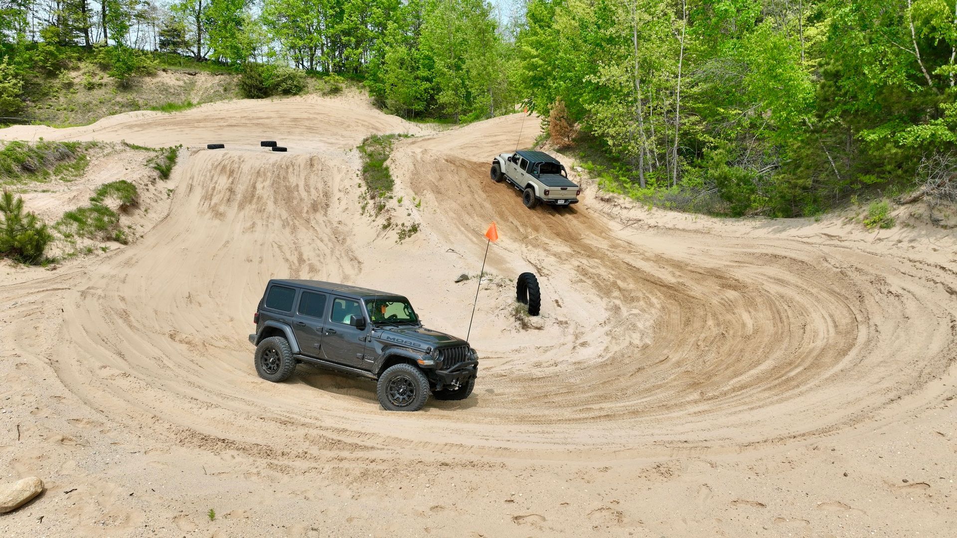 Jeep driving on sand course, other car descends ramp. Trees line the course.