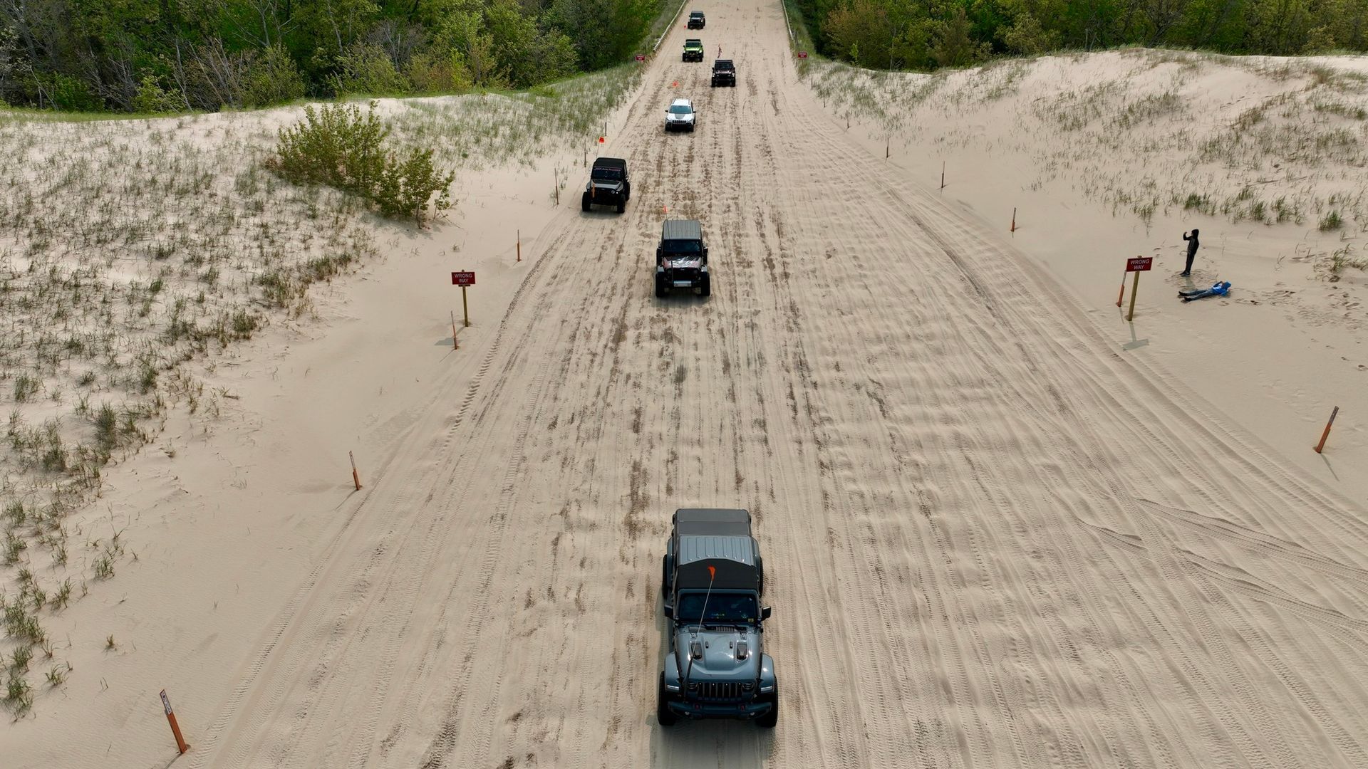 Jeeps driving on a sandy road, dunes on either side with green trees in the background.