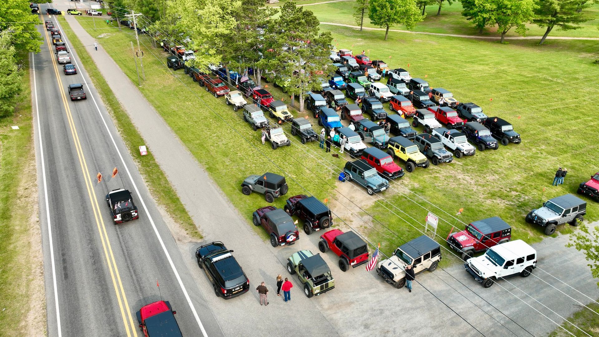 Cars lined up on grass and road, many Jeeps. People gather near vehicles.