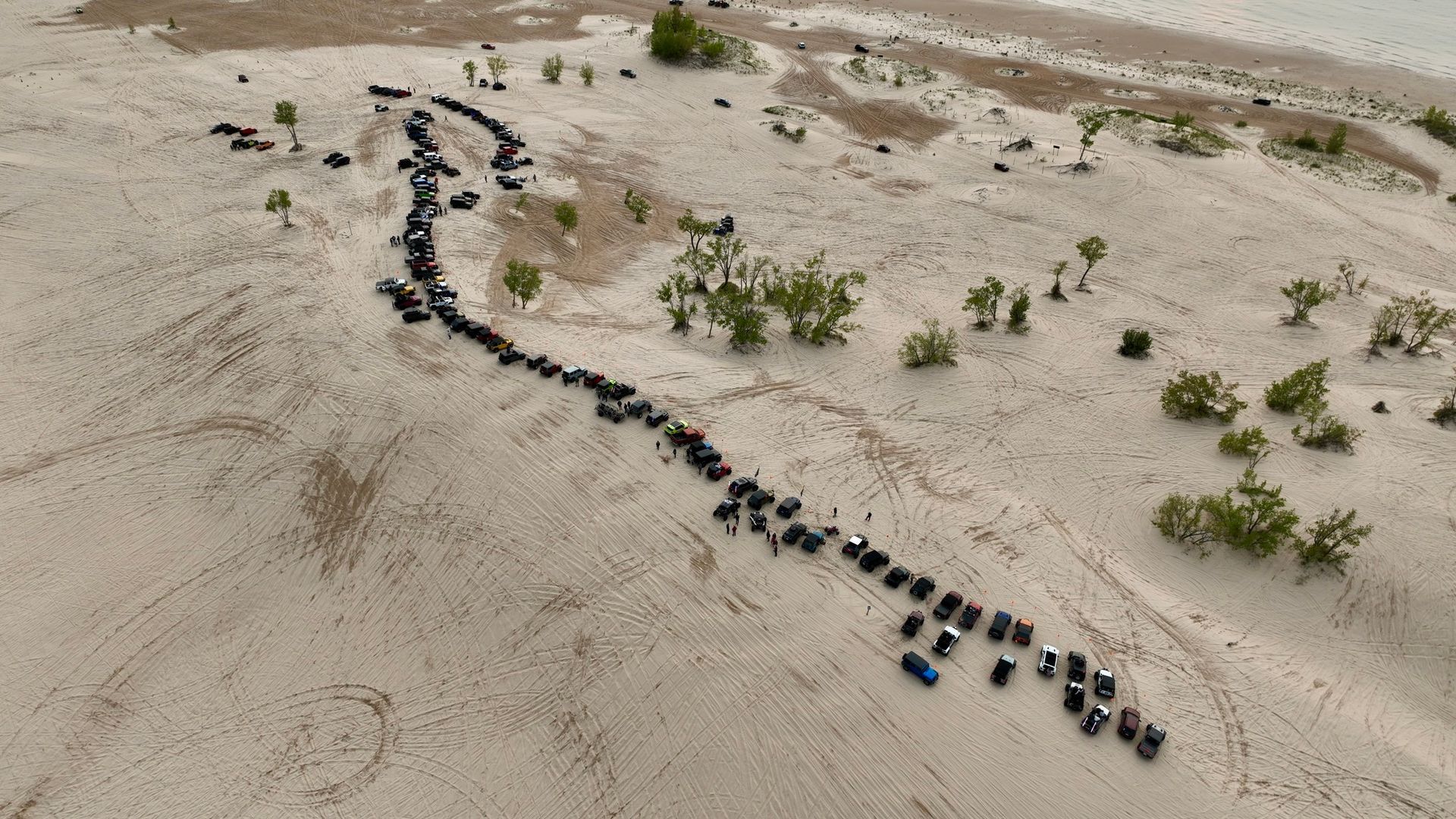 A long line of vehicles travels across a sandy landscape toward the horizon, possibly a beach or desert.