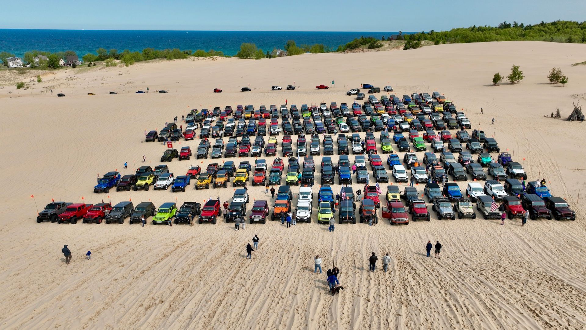 Many Jeeps parked on a sandy beach near a body of water, several people around.