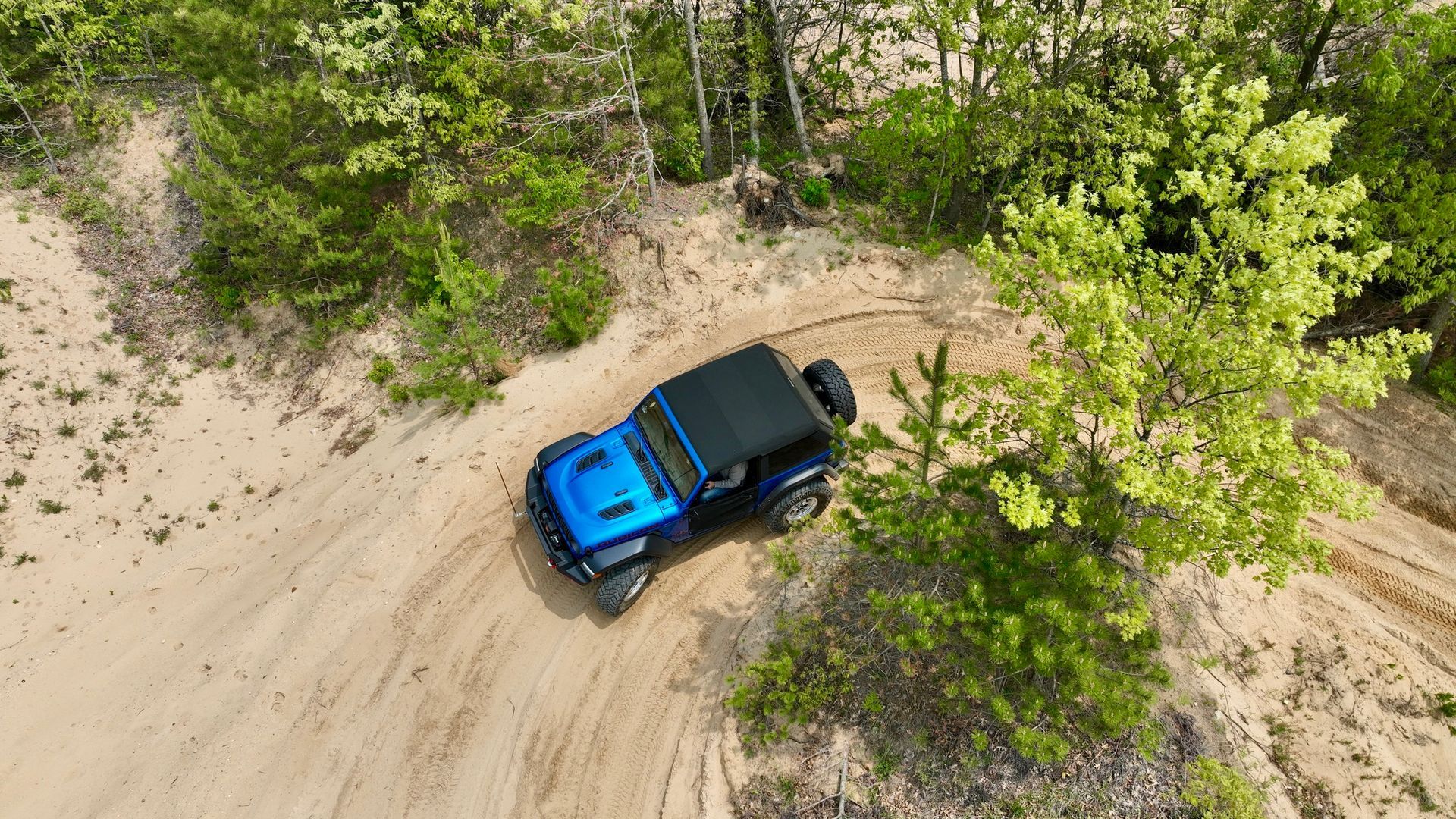 Blue Jeep driving on a sandy off-road trail surrounded by trees.
