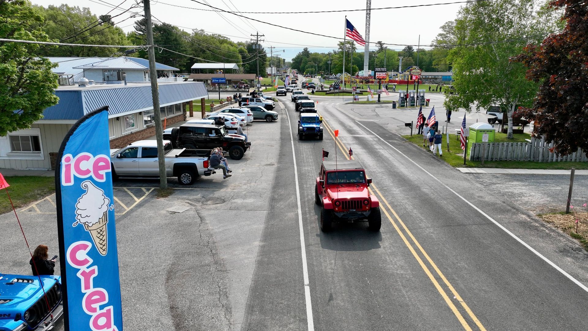 A parade with Jeeps and vehicles on a street, small town setting with ice cream sign and people watching.