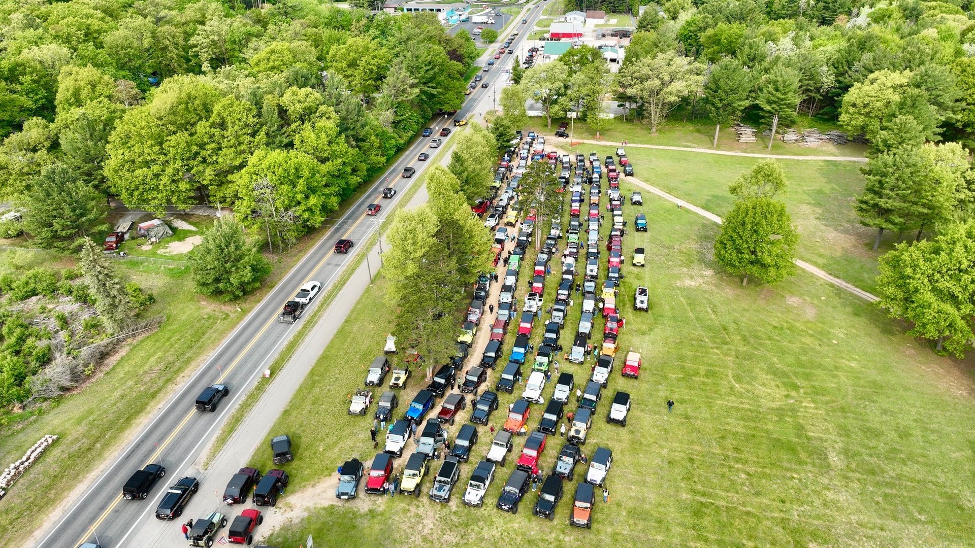 Aerial view of many parked Jeeps on grass, and cars on road, near trees.