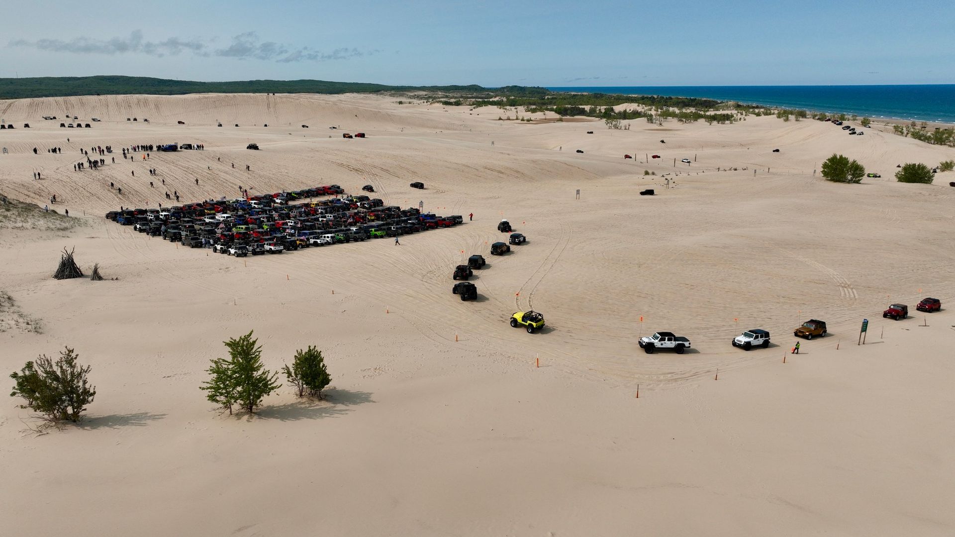Sandy dunes with a group of vehicles and people gathered, blue water in background.