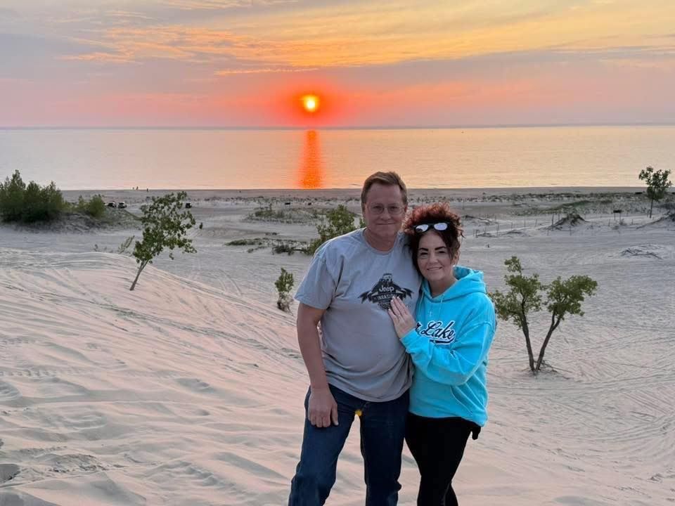 Couple on sand dune at sunset, orange sky, lake background.