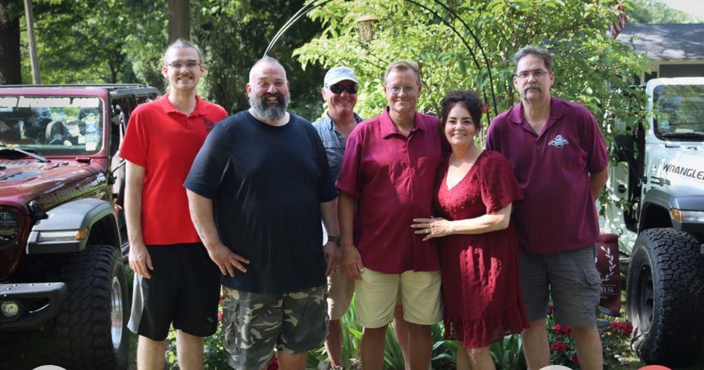 Group of people posing by two Jeeps, outdoors. Several are wearing red or purple shirts.