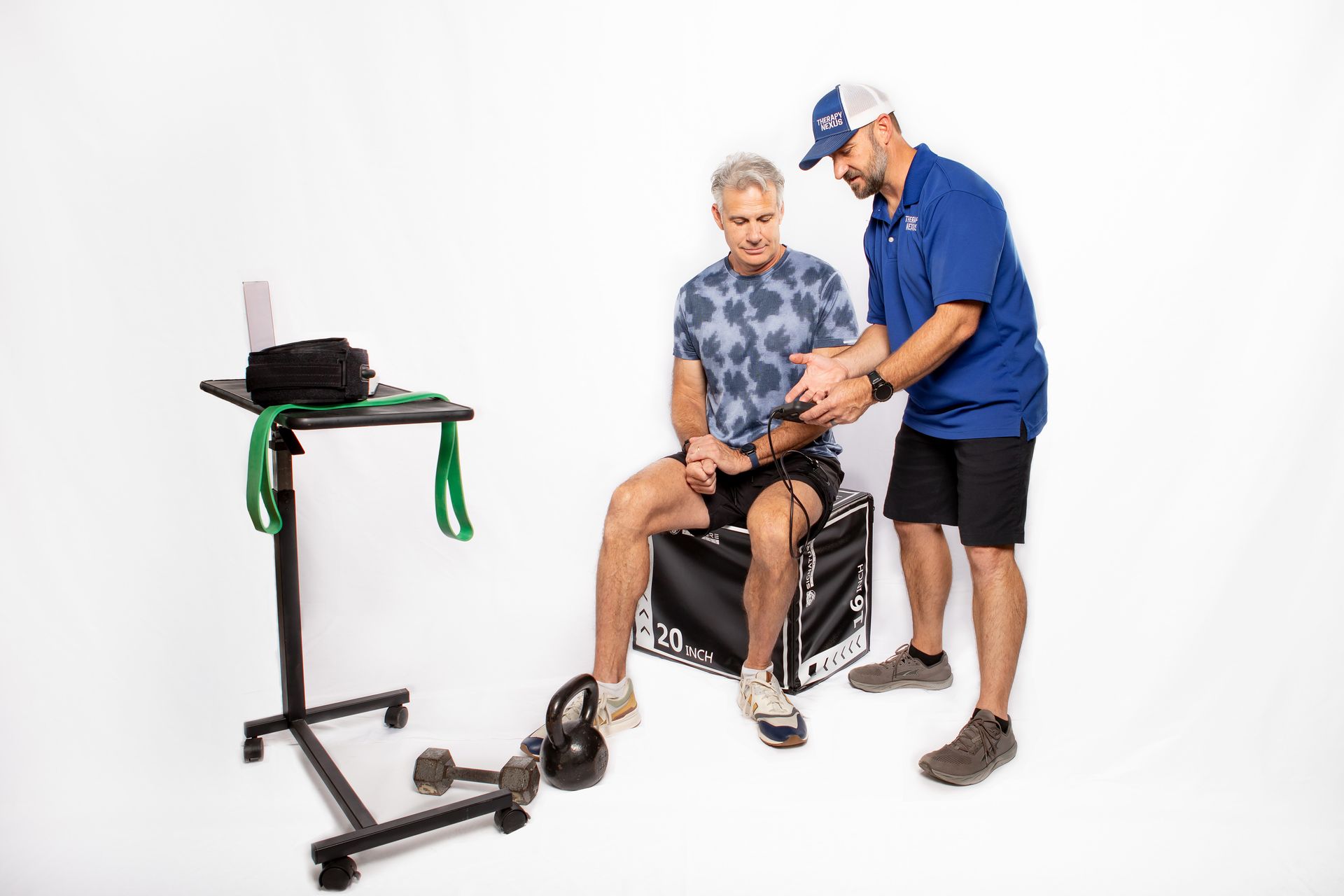 Man seated, trainer assisting with resistance band exercise. White background. Fitness equipment nearby.