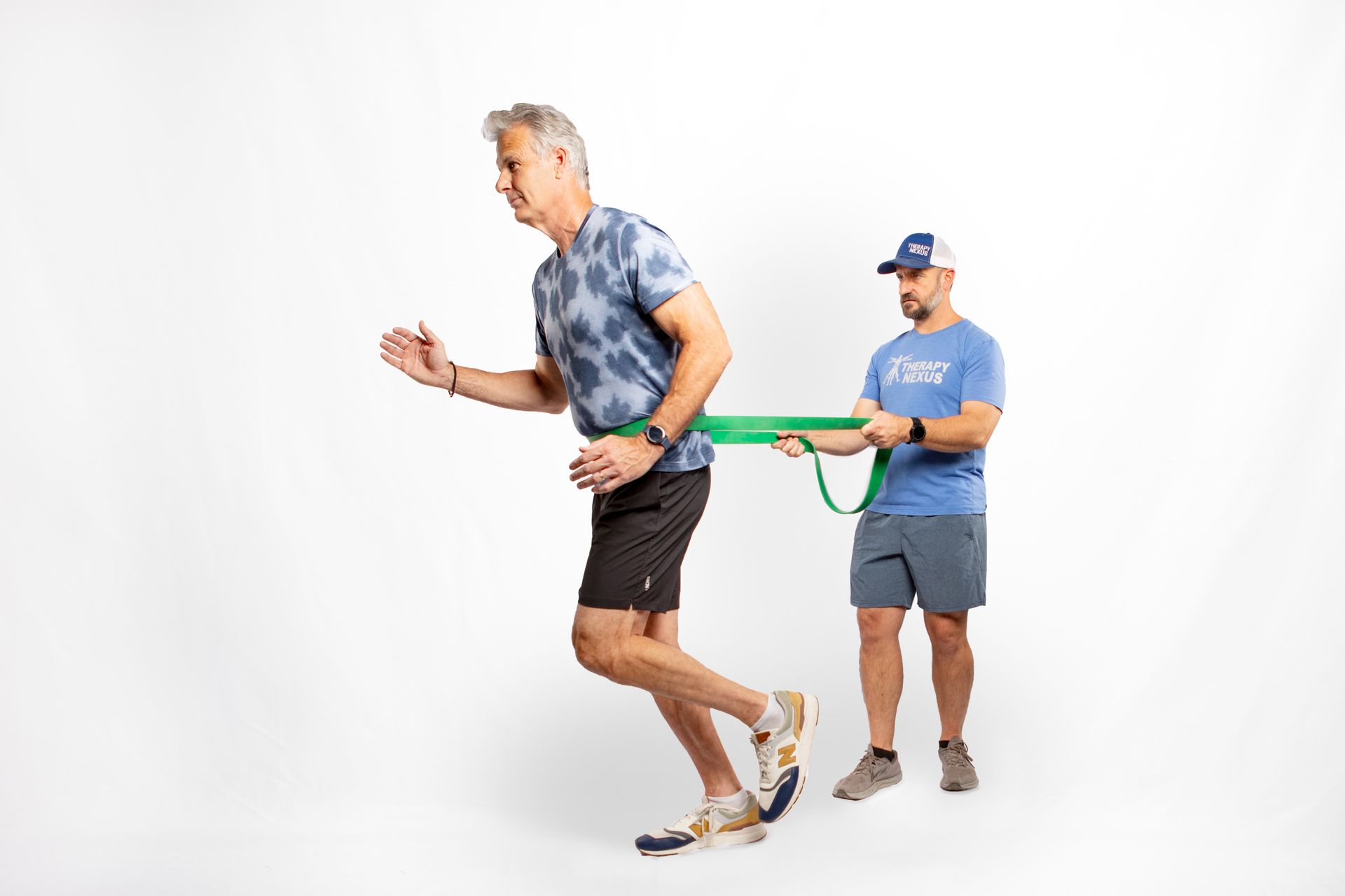 Man in activewear doing a resisted run with a green band held by another man in a gym setting.