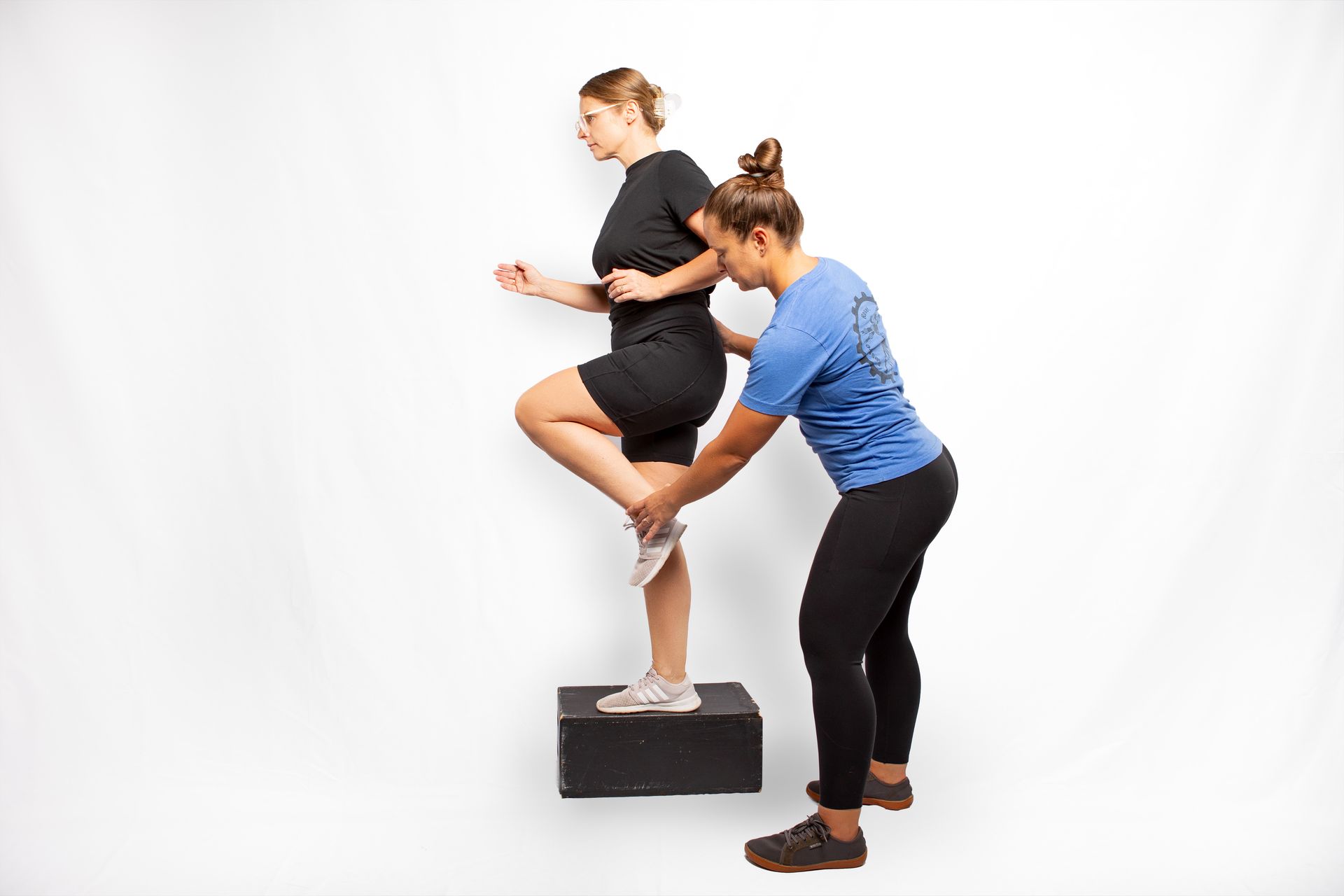 Woman stepping onto a box, assisted by another woman. They are in front of a white background.