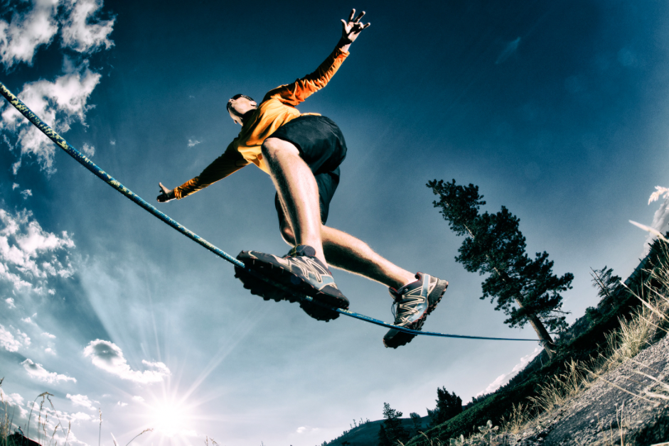 Person balances on a slackline outdoors, arms outstretched, blue sky, sunny.