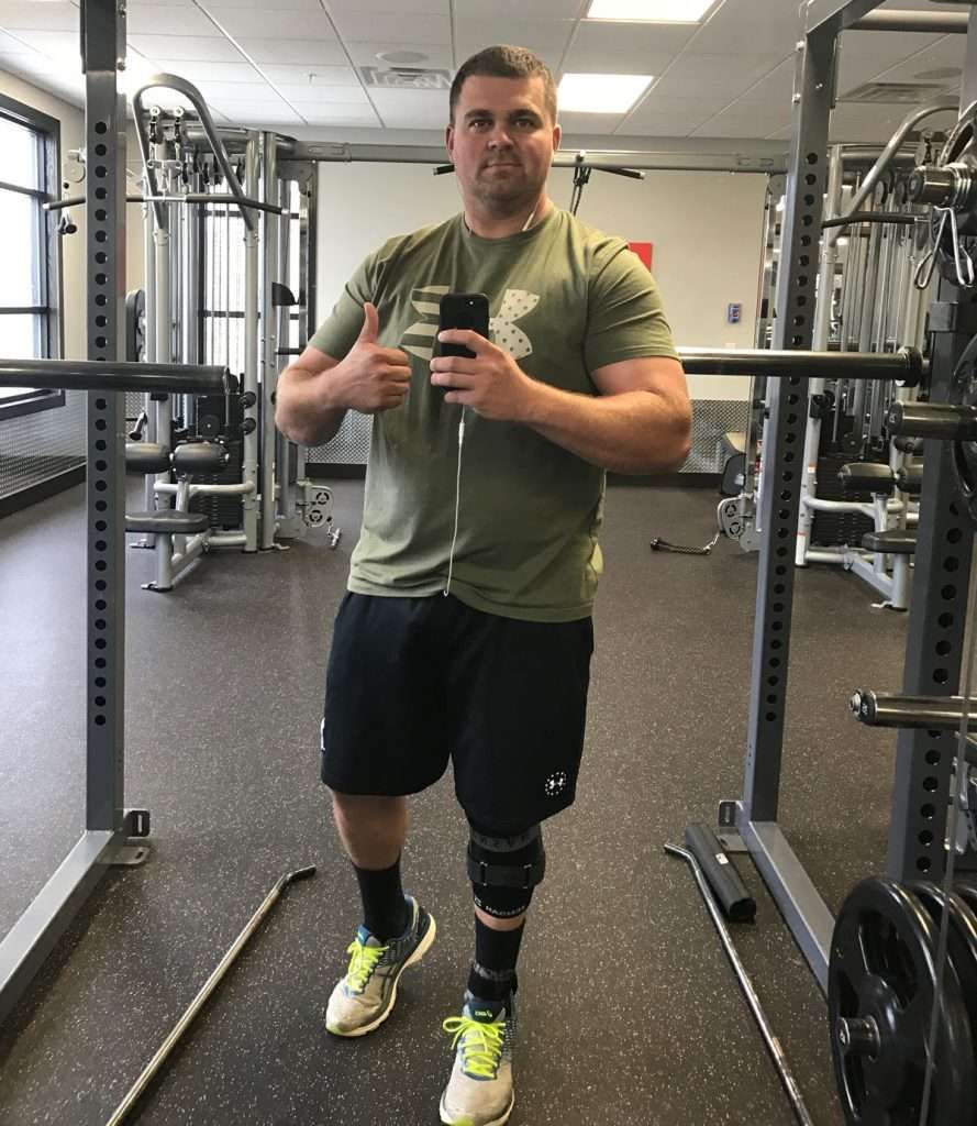Man with a prosthetic leg giving a thumbs up in a gym. He is wearing workout clothes and holding a phone.